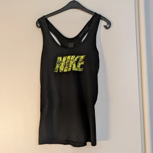 Nike Pro svart träningslinne M - Svart träningslinne från Nike Pro med gult ormmönstrat Nike-tryck framtill. Sportig racerback-modell i stretchigt material, perfekt för träning. Har Dri-FIT-tyg som andas. Snygg och funktionell för gymmet eller löprundan. Tyvärr materialetiketten avklippt, men i mycket bra skick! 