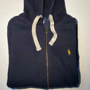 Ralph Lauren Zip Hoodie - Säljer nu denna snygga mörkblå Ralph Lauren Zip Hoodien med vita snören och ett gult märke. Nytt skick/mycket bra! Hör av dig om du undrar något, priset kan diskuteras!