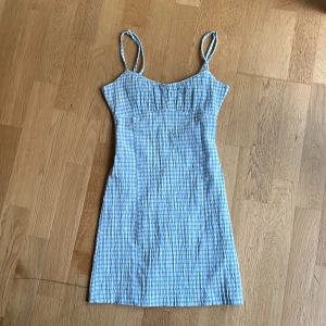 Blåvit rutig kortklänning Pull&Bear - Supersöt blåvit rutig kortklänning från Pull&Bear med smala axelband och figurnära passform. Klänningen har dragkedja i ryggen och är gjord i ett lätt bomullstyg, perfekt för varma dagar. Den har en klassisk somrig vibe och är enkel att styla. Bara använts en gång 💕
