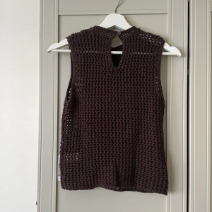 Brun virkad ärmlös topp med knappar - Säljer en brun, handvirkad ärmlös topp med rund hals och två knappar i nacken. Toppen har ett luftigt, grovmaskigt mönster. Ena knappen sitter aningen löst och fins en maska som har gått upp lite annars i bra skick! Använd 2 gånger❤️