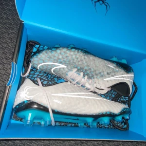 Nike Hypervenom Phantom fotbollsskor - Nike Hypervenom Phantom RGN SE FG i svart, grått och blått med coolt mönster och vita detaljer. Skorna har snörning, rund tå och platt sula, samt en spindelgrafik på hälen. Perfekta för dig som vill sticka ut på planen.