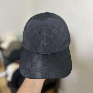 Svart Gucci keps med GG-mönster - Snygg svart keps från Gucci med klassiskt GG-monogram över hela framsidan. Kepsen har mesh på sidorna och bak, samt justerbar snapback. Materialet är en mix av polyester och bomull, vilket ger en lyxig känsla och bra passform och perfekt för sommaren🌤️