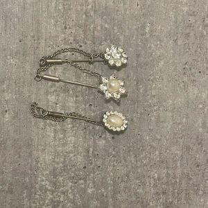 Tre silverfärgade broscher med pärlor - Säljer tre snygga broscher i silverfärg med kedja. Två av broscherna har vita pärlor i mitten och alla är dekorerade med glittriga stenar runt om. En brosch har blommig form, de andra är runda och ovala. Perfekt för att ge din outfit lite extra bling.