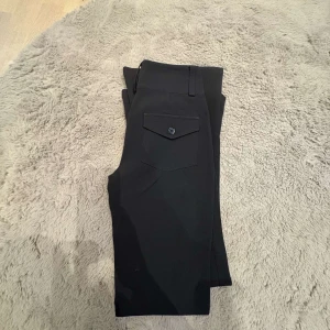 Svarta bootcut kostym byxor  - Svarta bootcut kostymbyxor med snygg ficka bak och klassisk passform. Byxorna har en stilren look och är perfekta för dig som gillar en tidlös vibe. Materialet känns mjukt och stretchigt, vilket gör dem bekväma hela dagen. Aldrig använda 