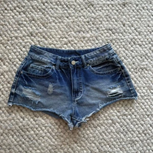 Blå låga jeansshorts - Snygga blå jeansshorts med låg midja och slitna detaljer framtill. Shortsen har råa kanter och klassiska fickor både fram och bak med coola knappar på bakfickorna. Perfekta för sommardagar när du vill ha en avslappnad och trendig look.