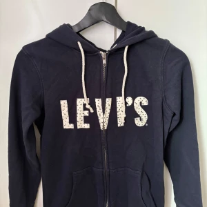 Mörkblå hoodie från Levi's med dragkedja - Marinblå hoodie från Levi's med vit logga framtill dekorerad med små svarta detaljer. Tröjan har huva med vita snören, dragkedja och två fickor framtill. Tillverkad i mjuk bomull och har långa ärmar. 