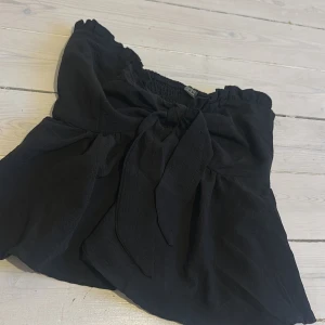 Svart offshoulder blus med knyt - Supersöt svart offshoulder blus med volangig nederdel och bred smock upptill. Framtill finns en stor knytrosett som ger en extra detalj. Perfekt att styla med jeans eller kjol för en trendig look.