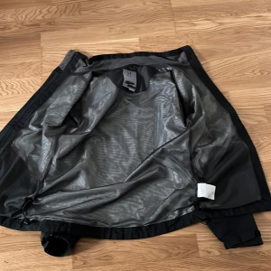 Svart vindjacka från Haglöfs - Snygg svart vindjacka från Haglöfs med huva och dragkedja framtill. Jackan har reflekterande detaljer på ärmarna, två stora fickor och justerbara muddar. Perfekt för blåsiga dagar och outdoor-äventyr. Materialet är lätt och känns smidigt.