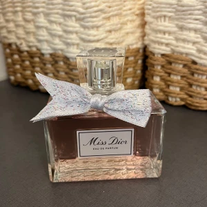 Miss Dior Eau de Parfum 100ml - Miss Dior Eau de Parfum i en lyxig 100ml glasflaska med ljusrosa doftvätska. Flaskan har en elegant fyrkantig form och pryds av en glittrig, ljusgrå rosett vid halsen. Klassisk och feminin design från Dior, perfekt för dig som älskar exklusiva parfymer. Aldrig använd helt ny! 