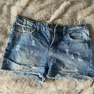 Blå slitna jeansshorts Sunny Star - Snygga blå lågmidjade jeansshorts från Sunny Star med slitna detaljer och fransig kant. Klassisk femficksmodell med dragkedja och knapp. Perfekta för varma dagar när du vill ha en avslappnad och cool stil.