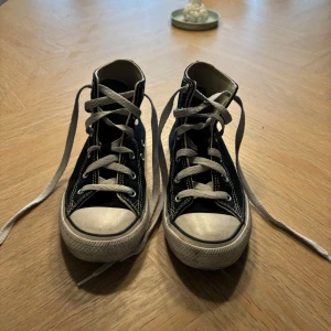 Svarta Converse Chuck Taylor All Star - Klassiska svarta Converse Chuck Taylor All Star high tops med vita snören och gummisula. Skorna har rund tå, tydliga vita sömmar och den ikoniska loggan på sidan. Tillverkade i canvas med vita detaljer och silverfärgade öljetter.