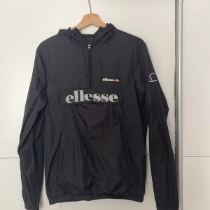 Svart vindjacka från Ellesse - Svart vindjacka från Ellesse med stor logga framtill och mindre logga på bröstet. Jackan har huva med snörning, halv dragkedja och elastiska muddar. Perfekt för dig som gillar sportig och clean stil. Lätt och smidig att bära, passar till många outfits.