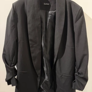 Svart kavaj från Boohoo - Stilren svart kavaj från Boohoo i storlek M. Kavajen har klassisk passform med breda slag och två fickor framtill. . Perfekt att slänga över en t-shirt eller skjorta för en snygg och classy look.