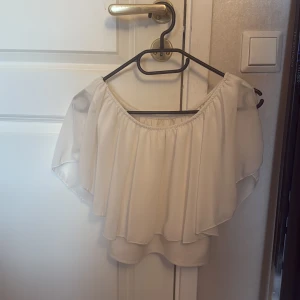 Vit offshoulder blus med volang - Superfin vit blus med offshoulder-modell och dubbla lager volang. Tunn och luftig, perfekt för varma dagar. Resår upptill för snygg passform och flowig känsla. Passar till både jeans och kjol för en trendig look.