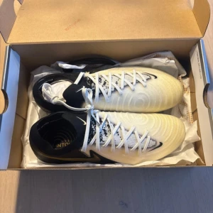 Nike Phantom GX II Elite FG fotbollsskor - Nike Phantom GX II Elite FG i vitt, svart och guld. Designade för gräsplan med greppig sula och låg profil. Helt nya och helt oanvända! 