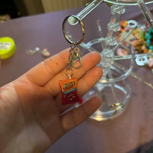 Nyckelring med Cheetos-charm - Cool nyckelring med en miniatyrpåse Cheetos som hänge. Färgglad design i rött och orange med blå detaljer, perfekt för dig som gillar snacks och vill ha något unikt på väskan eller nycklarna.