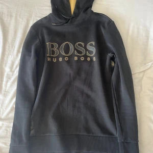 Svart hoodie från Hugo Boss - Svart hoodie från Hugo Boss med stor BOSS-logga i guld på bröstet och guldfärgad insida på huvan. Tröjan har långa ärmar, ribbade muddar och snörning i huvan. Snygga BOSS-detaljer längs sidorna. Perfekt för en stilren och avslappnad look. Storlek S.