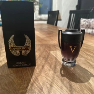 Herrparfym 100ml - Parfymen Inversion Eau de Parfum kommer i en svart, stilren box med guld- och silverdetaljer och vingar. Flaskan är formad som en pokal i mörkt glas med svarta handtag och ett stort V i guld på framsidan. Rymmer 100 ml och har en exklusiv vibe. (Ny, vet inte om äkta, oanvänd Invictica Victory)