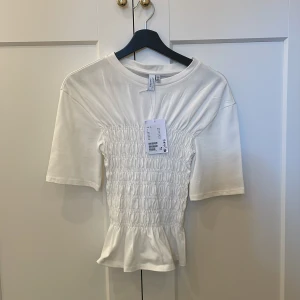Vit smockad topp från & Other Stories - Superfin vit topp från & Other Stories med smockad midja och lätt volang nertill. Gjord i 100% bomull, rund hals och korta ärmar. Perfekt för dig som gillar en feminin och trendig look med snygga detaljer. Den är aldrig använd och bara hängt i min garderob. Nypris 390kr