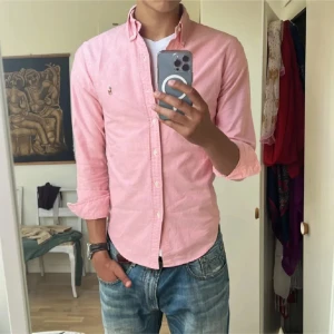 Ralph Lauren Skjorta (NY SKICK) - Säljer en klassisk rosa skjorta från Ralph Lauren i SLIM FIT med button-down krage och broderad logga på bröstet. Skjortan har lång ärm, normal passform och är tillverkad i mjuk bomull. Perfekt för dig som gillar stilrena och tidlösa plagg.