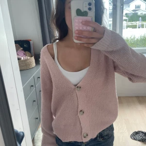 Ljusrosa stickad kofta med knappar - Supermjuk och mysig ljusrosa stickad kofta med v-ringning och tre stora knappar framtill. Koftan har lång ärm och en lite loose passform, perfekt att slänga över ett linne eller t-shirt. Enkel och stilren design som är lätt att matcha.