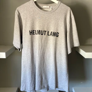 Grå t-shirt från Helmut Lang - Snygg grå t-shirt från Helmut Lang med svart logotyptryck på bröstet. Klassisk rund halsringning och avslappnad passform. Perfekt för dig som gillar minimalistisk och stilren design.