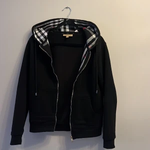 Svart Burberry hoodie - Svart hoodie från Burberry med dragkedja och klassiskt rutigt foder i huvan. Jackan har långa ärmar, fickor framtill och snygga detaljer som silverfärgad dragkedja. Perfekt för dig som gillar stilren design med en touch av lyx. (Något tajtare i midjan och ska passa en ”XS”)