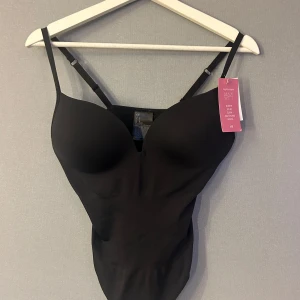 Svart body från H&M - Svart body från H&M med vadderade kupor och djup v-ringning. Topp med smala, justerbara axelband och slät, tight passform som formar kroppen. Perfekt att styla under kläder. Materialet är stretchigt och följsamt för en snygg siluett.
