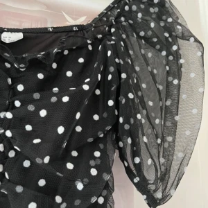 Svart prickig meshklänning - Supersöt svart kortklänning i mesh med vita prickar och puffiga kortärmar. Klänningen har rynkade detaljer framtill och en luftig känsla tack vare det tunna materialet. 