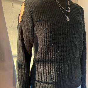 Svart stickad tröja med öppna axlar - Snygg svart stickad tröja med unika öppna axlar. Perfekt för att ge en stilren look med en twist. Tröjan har långa ärmar och en ribbad struktur som ger en skön passform.