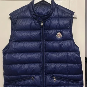 Blå dunväst från Moncler - Snygg blå dunväst från Moncler med dragkedja och två praktiska fickor framtill. Perfekt för kyligare dagar när du vill hålla stilen. Moncler-loggan pryder bröstet för en exklusiv touch. Sitter bra på mig som är 185cm