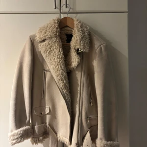 Beige jacka med fuskpäls - Säljer en snygg beige jacka med fuskpälsdetaljer. Jackan har dragkedja och bälte, vilket ger en cool look. Perfekt för kyligare dagar med sin varma och stiliga design.