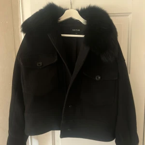 Svart jacka från Meotine - Snygg svart jacka från Meotine med en lyxig pälskrage. Jackan har knappar framtill och två fickor med lock. Materialet består av en blandning av ull och polyester, vilket gör den både varm och stilren. Lite nipprig på vissa ställen men inget man märker av! Köpt för 3500kr, säljer för 1800kr.