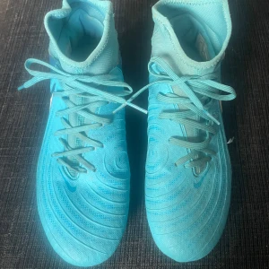 Blå Nike Phantom Luna fotbollsskor - Snygga blå Nike Phantom Luna fotbollsskor med snörning och rund tå. Perfekta för fotbollsplanen med sin stilrena design och funktionella dobbar. Skorna har en iögonfallande blå färg med vit Nike-logga på sidan.