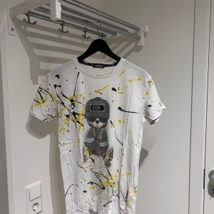 Vit t-shirt med färgstänk och tryck icon - Cool vit t-shirt från Icon med färgstänk i gult och svart. Framsidan har ett tryck av en katt med mössa och solglasögon. Perfekt för en avslappnad stil.