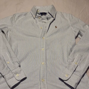 Randig skjorta från Ralph Lauren - Snygg blå- och vitrandig skjorta från Ralph Lauren med broderad logotyp på bröstet. Skjortan har långa ärmar och knappar framtill. Perfekt för en stilren look. Pris kan diskuteras!!!