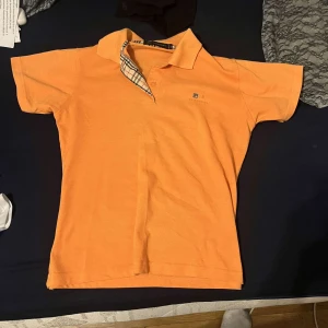 Orange pikétröja från Burberry - Snygg orange pikétröja från Burberry med klassisk krage och korta ärmar. Tröjan har ett diskret märke på bröstet och ett stilrent rutigt mönster på insidan av kragen. Perfekt för en avslappnad men stilren look. Priser kan diskuteras