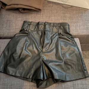 Svarta skinnshorts - Snygga svarta shorts i skinnimitation med hög midja och bred resår. Perfekta för en trendig look, med stilrena fickor bak och en elegant passform.