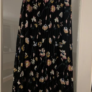 Blommig långkjol från Carin Wester - Säljer en vacker långkjol från Carin Wester med ett blommigt mönster i rosa, gult och vitt på en svart bakgrund. Kjolen har en flödande passform och är perfekt för både vardag och speciella tillfällen. 