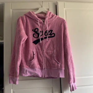 Rosa hoodie från Svea - Mysig rosa hoodie från Svea med dragkedja och huva. Framsidan har en stor svart text och baksidan pryds av siffrorna '98'. Perfekt för en avslappnad stil med sina långa ärmar och mjuka material.