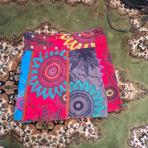 Färgglad kortkjol med mönster - Säljer en unik och färgglad kortkjol med ett mixat mönster i rött, blått och turkos. Kjolen har knappar i midjan och ett patchwork-design med cirkulära och blommiga motiv. Perfekt för dig som vill sticka ut!