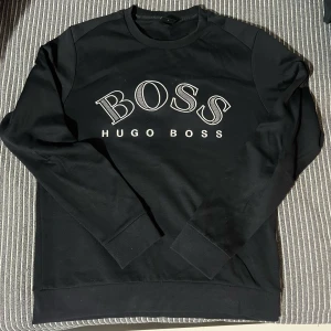 Svart tröja från Hugo Boss - Snygg svart tröja från Hugo Boss med blått tryck på bröstet. Perfekt för en stilren look. Tröjan har långa ärmar och rund halsringning.