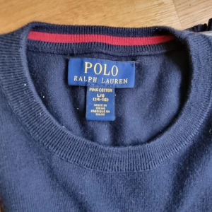 Mörkblå tröja från Ralph Lauren - Säljer en stilren mörkblå tröja från Ralph Lauren i pima bomull. Tröjan har en broderad röd logotyp på bröstet och ribbade muddar vid ärmslut och nederkant. Perfekt för en klassisk look.