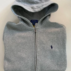 Ralph Lauren Hoodie Grå  - Ralph Lauren hoodie – Grå – Strl L (14–16 år / ca 170 cm)  Klassisk grå hoodie från Ralph Lauren i riktigt fint skick. Stilren, mjuk och bekväm – passar perfekt till både vardag och chill. 	•	Storlek: L (14–16 år / ca 170 cm) 	•	Färg: Grå 	•	Väldigt fint skick, inga fläckar eller hål 	•	Original – klassisk logga framtill