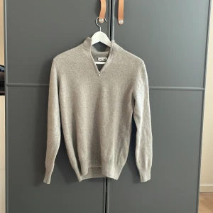 Grå kashmirtröja från Soft Goat - Säljer en stilren grå kashmirtröja från Soft Goat med dragkedja vid halsen. Tröjan har långa ärmar och en ribbad krage samt muddar. Perfekt för kyliga dagar!
