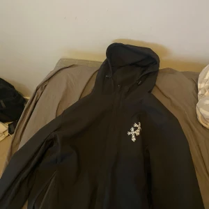 Svart jacka med korsdetalj - Säljer en svart jacka med en stor vit korsdetalj på ryggen. Jackan har en huva och dragkedja framtill. Perfekt för en stilren look.