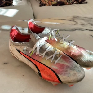 Flerfärgade fotbollsskor från Puma - Säljer ett par lätta och färgglada fotbollsskor från Puma. Skorna har en vit bas med inslag av rött, orange och svart. De är designade med snörning och har en strömlinjeformad siluett för optimal prestanda på planen. Bara lite tuggummi under skon som går att pilla bort lätt 