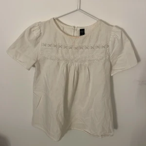 Vit blus med spetsdetaljer - Säljer en blus med korta ärmar och vackra spetsdetaljer på framsidan. Blusen har en rund halsringning och knäppning bak. Perfekt för en somrig dag!