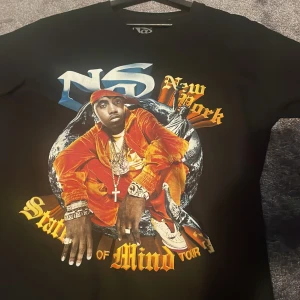 Svart hip hop  Nas T-shirt  - Säljer en svart t-shirt med ett coolt tryck av Nas i rött och gult. Perfekt för fans av hiphop och streetwear. Tillverkad i 100% ekologisk bomull för en skön känsla. Passar perfekt till vardags eller konsert.