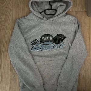 Grå hoodie från Trapstar - Säljer en grå hoodie från Trapstar med ett tryck av en panter och texten 'London Shooters' på framsidan. Hoodien har en klassisk design med känguruficka och justerbar huva. Perfekt för en avslappnad stil.